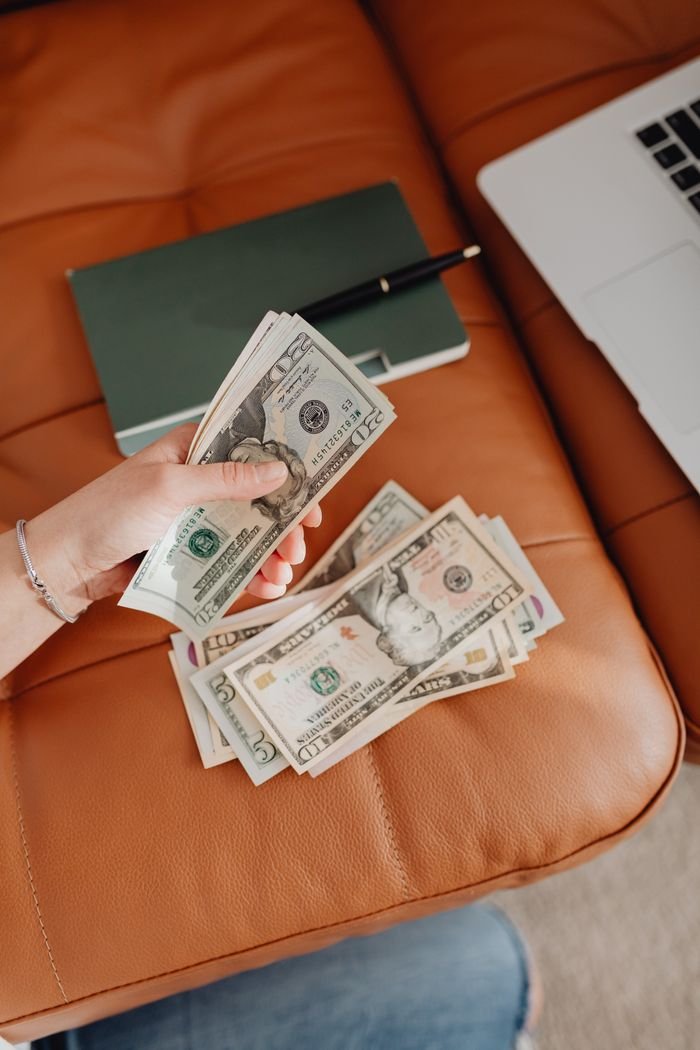 Person counting US dollars on a leather couch with a notebook and laptop nearby.